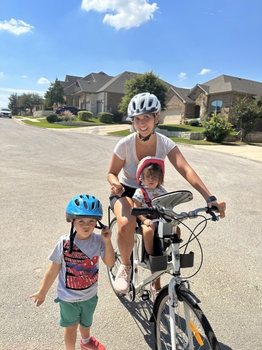 The author is on her bike with her baby on the baby seat in front of her. Her son is standing next to her and all three are wearing bike helmets.