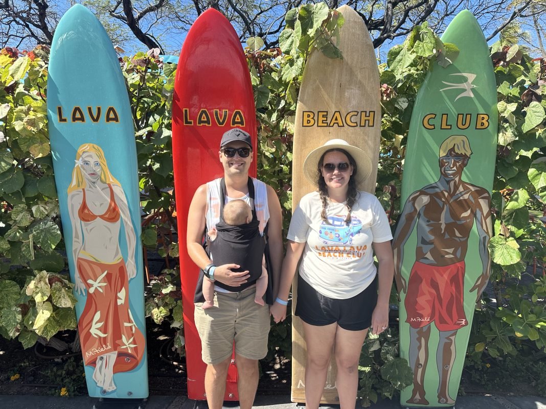 Sophie, a brown haired woman stands holding her baby wearing husbands hand in front of four surf boards which read lava lava beach club