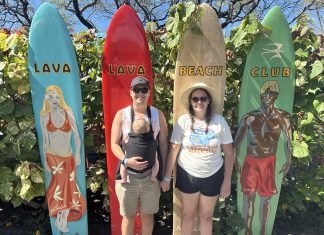 Sophie, a brown haired woman stands holding her baby wearing husbands hand in front of four surf boards which read lava lava beach club